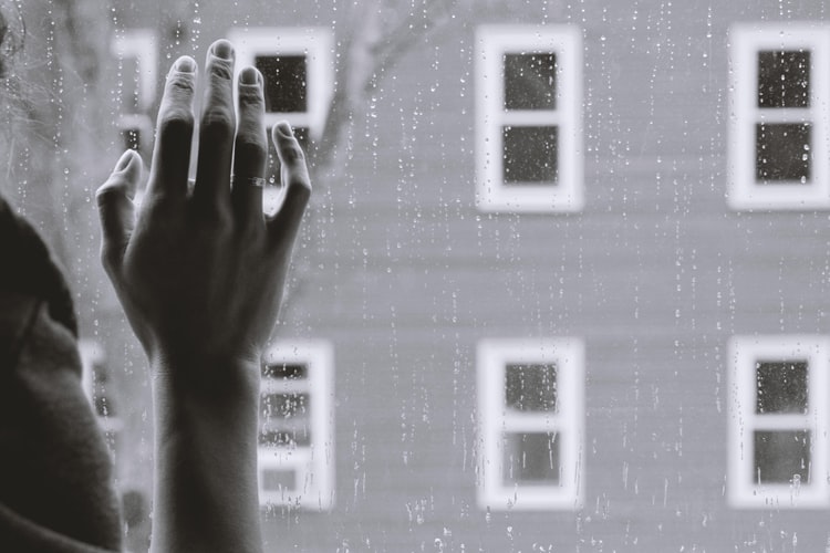 A greyscale photo of hand touching a window glass with rain drops on the exterior side