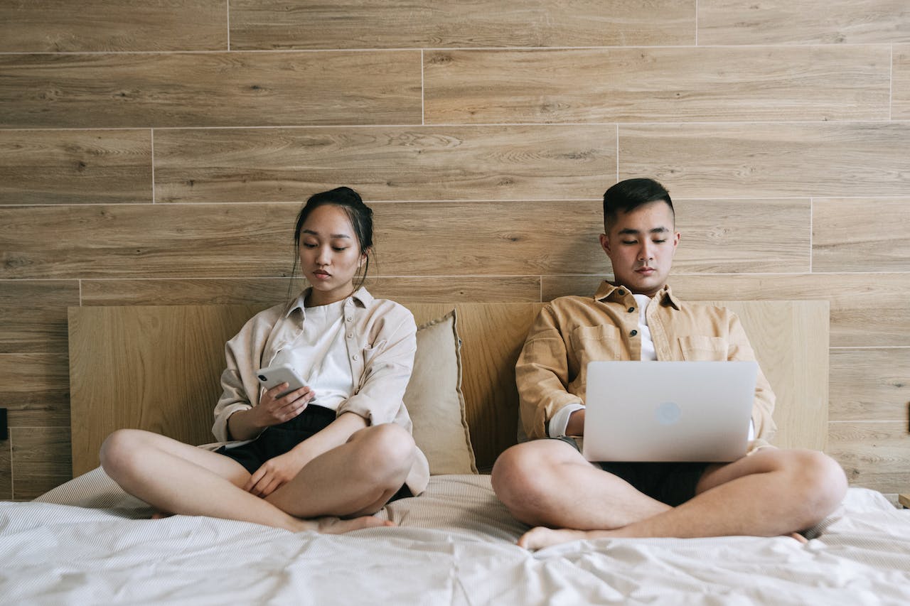 A young asian couple sitting in bed, the male using a laptop, the female using her mobile phone.