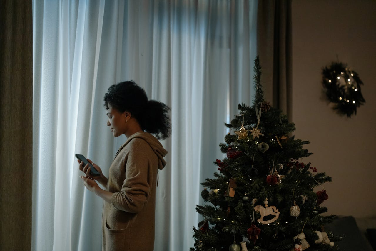 Woman holding a phone with both hands, Christmas tree behind her and a window in front.